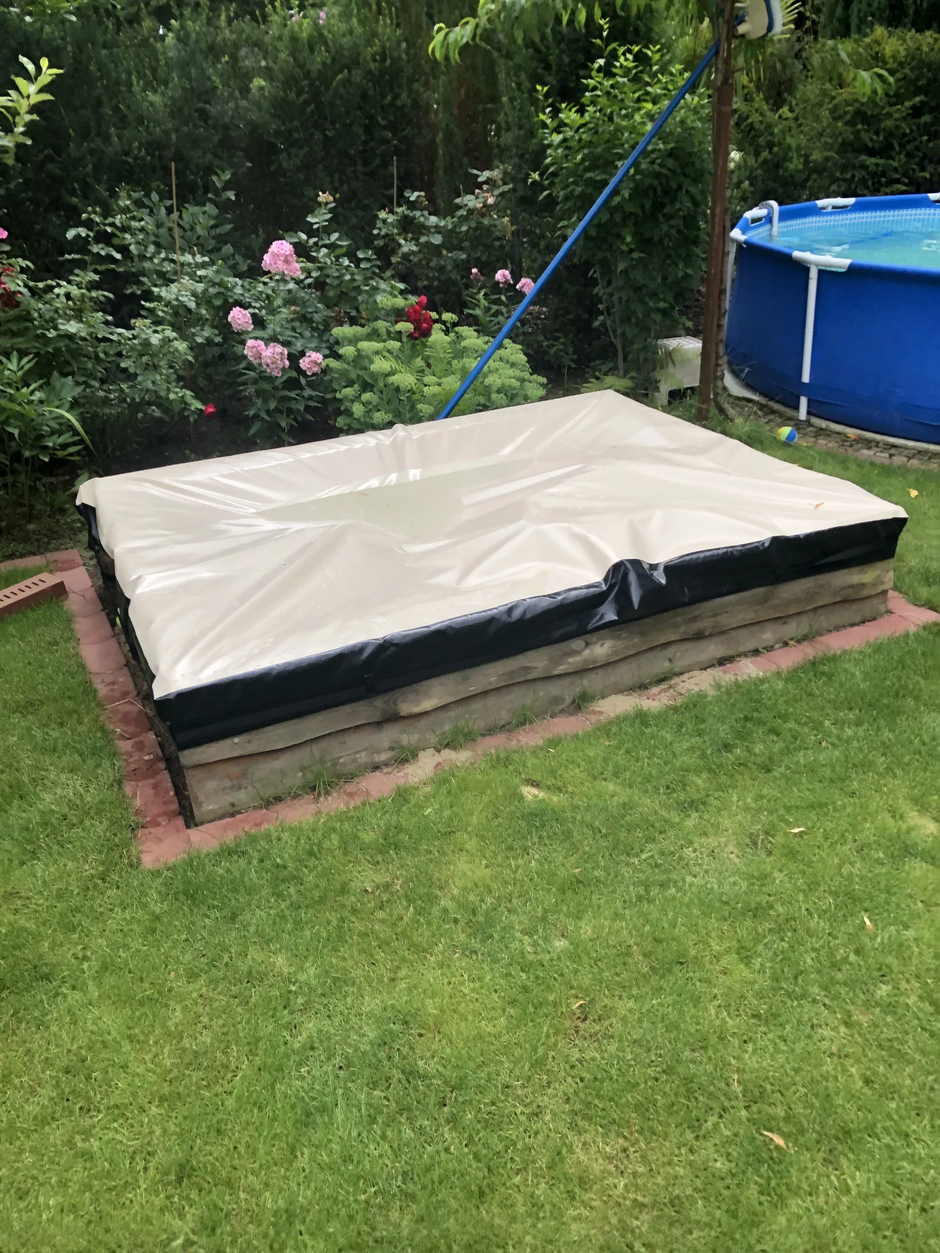 Rechteckige beige Abdeckung mit dunklem Rand auf einer erhöhten Liegefläche im Garten.